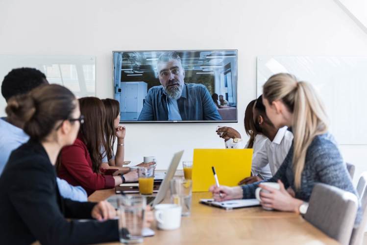 Office workers in a boardroom talking to a remote employee through an interactive display.