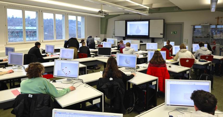 Students following a lesson on their laptops. An interactive whiteboard is visible in the background.